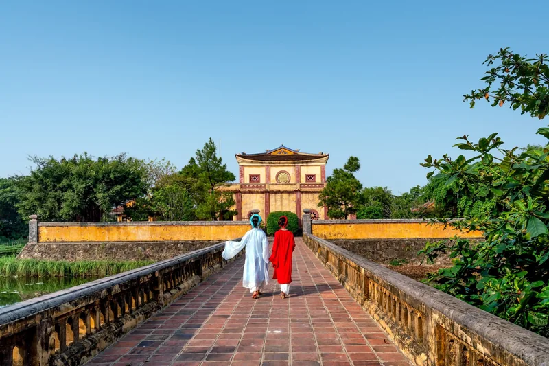 Hai cô gái mặc áo dài nhiều màu bước qua cầu gạch hướng tới cổng thành cổ ở Huế
