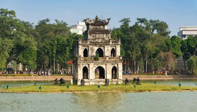 Tháp Rùa đứng trên gò đất nhỏ giữa hồ Hoàn Kiếm, Hà Nội, bao quanh bởi cây xanh và người dân dạo bước ven hồ.