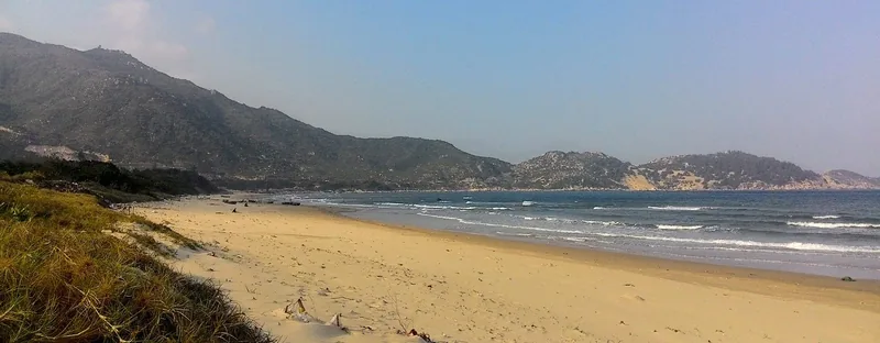 Bãi biển cát rộng trải dài dọc bờ biển gần bán đảo Phương Mai ở Quy Nhơn, với những ngọn đồi xanh phía xa.