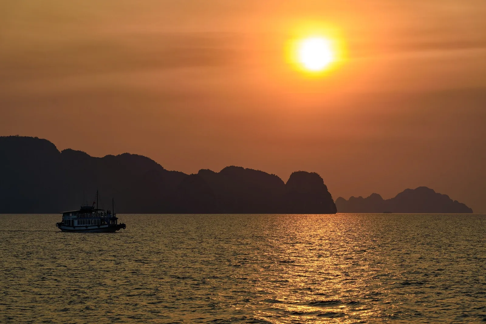Một thuyền du lịch lướt qua các đảo đá vôi in bóng dưới ánh hoàng hôn rực cam ở Vịnh Hạ Long.