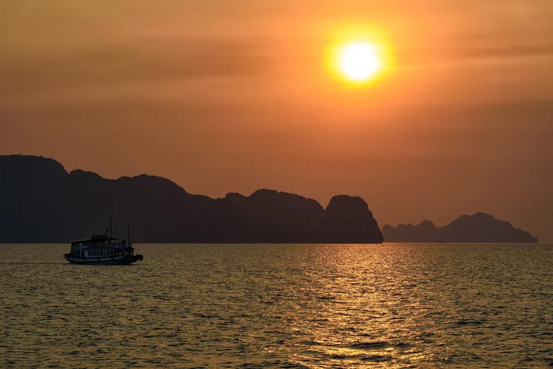 Một thuyền du lịch lướt qua các đảo đá vôi in bóng dưới ánh hoàng hôn rực cam ở Vịnh Hạ Long.